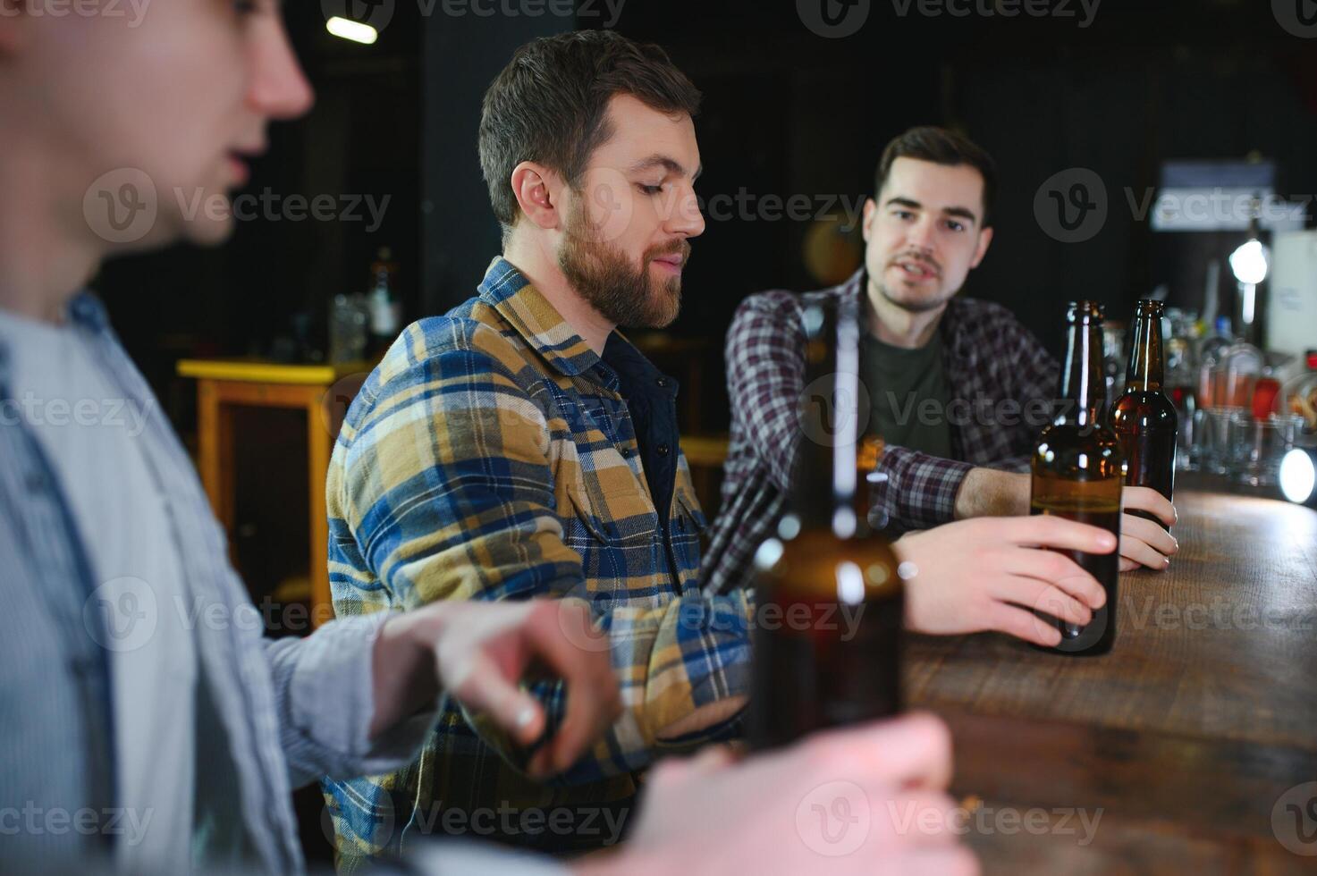 Three young men in casual clothes are smiling, holding bottles of beer while standing near bar counter in pub photo