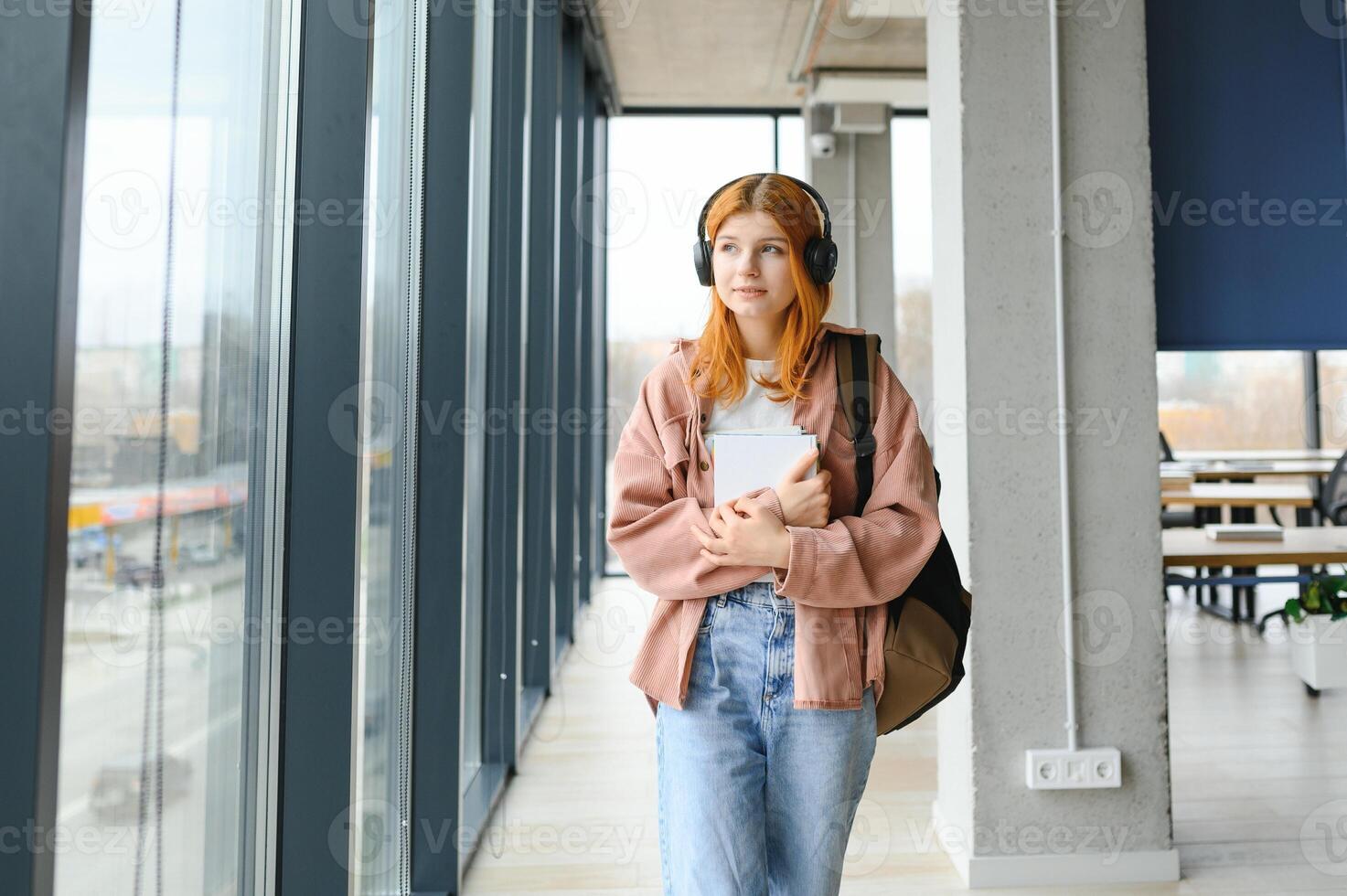 Red-haired student girl stands by the window with books and a backpack photo