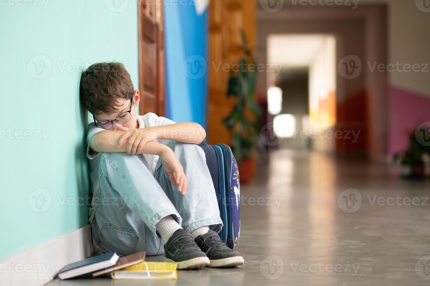 Little boy sitting alone on floor after suffering an act of bullying. Sad young schoolboy sitting on corridor with hands on knees and head between his legs photo