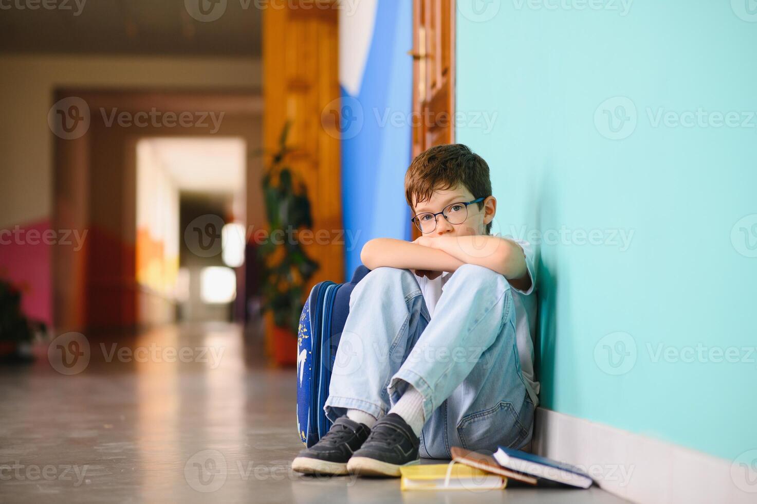 Little boy sitting alone on floor after suffering an act of bullying. Sad young schoolboy sitting on corridor with hands on knees and head between his legs photo