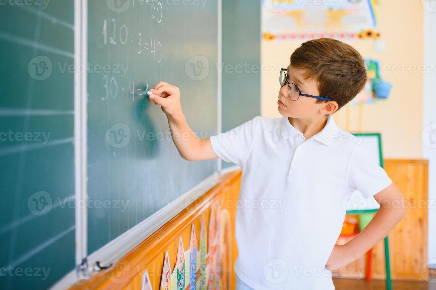 Cute little schoolboy diligent write chalk solve math problem dressed stylish uniform classroom blackboard background photo