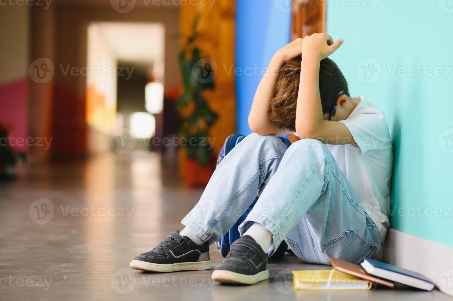 Little boy sitting alone on floor after suffering an act of bullying. Sad young schoolboy sitting on corridor with hands on knees and head between his legs photo