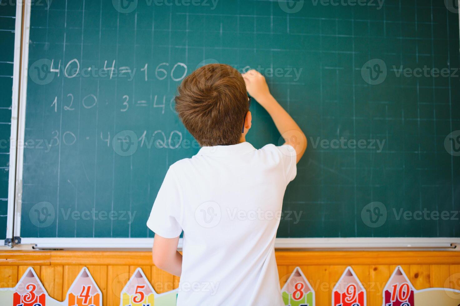 Cute little schoolboy diligent write chalk solve math problem dressed stylish uniform classroom blackboard background photo