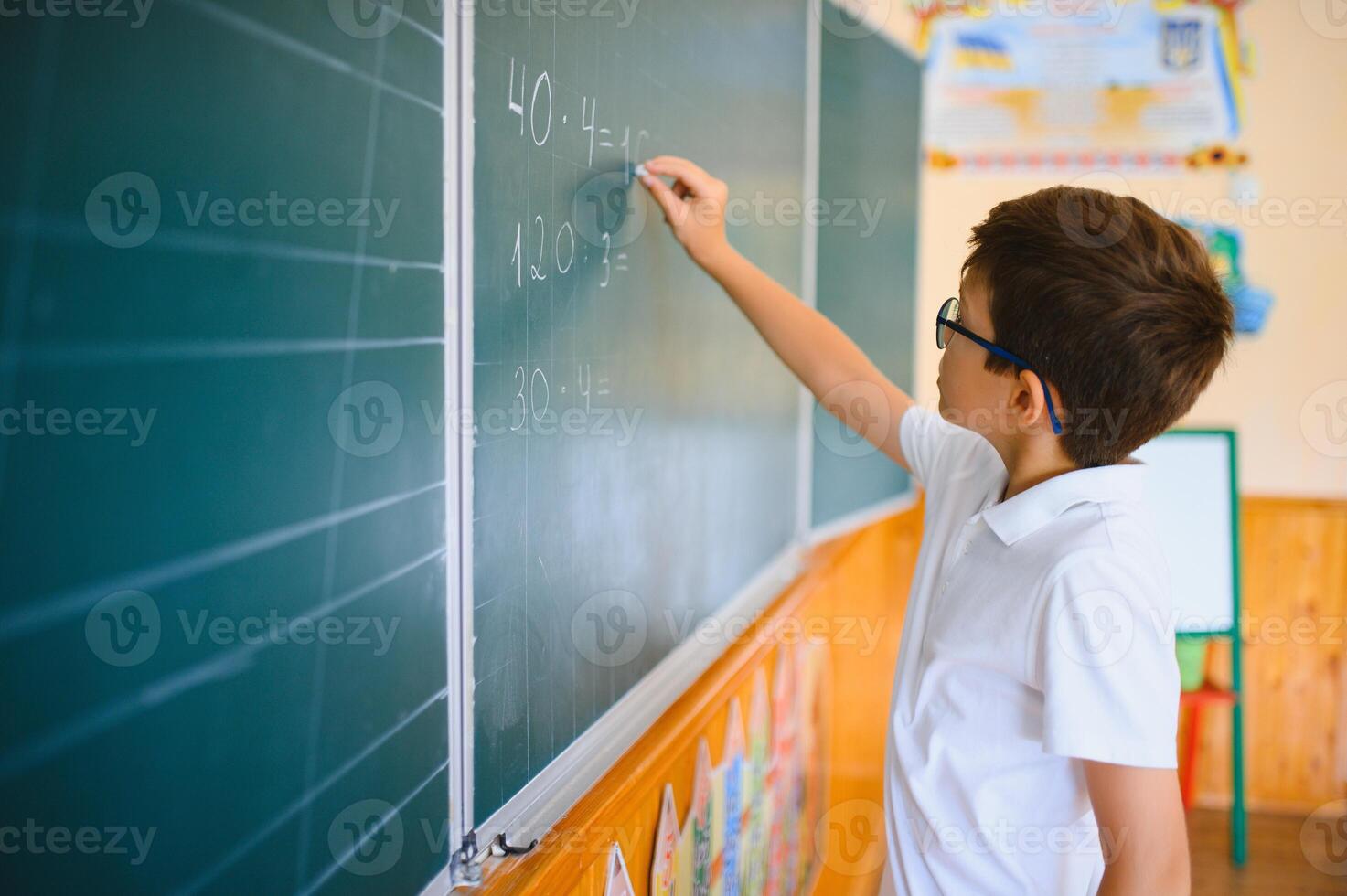 Cute little schoolboy diligent write chalk solve math problem dressed stylish uniform classroom blackboard background photo