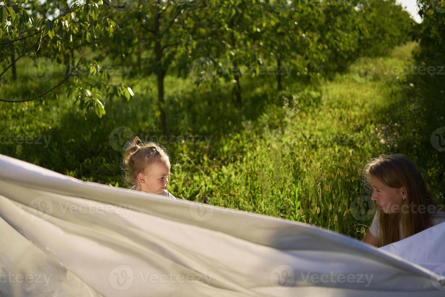 a family of four is playing with a white sheet in the garden, a toddler wants to flutter the sheet photo