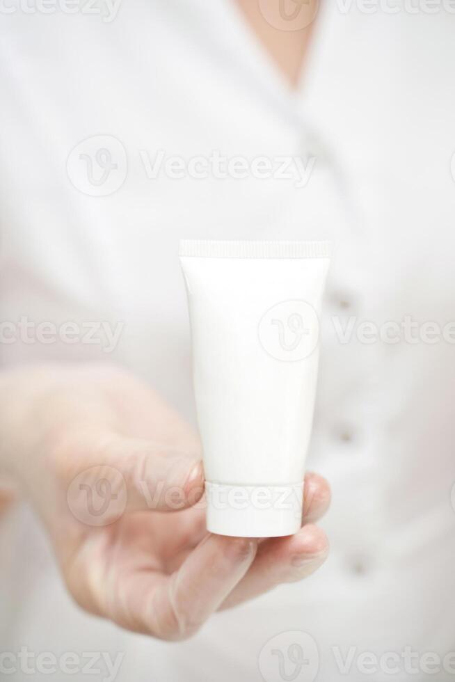 Hand in glove holding empty facial cream product. Mockup cosmetics bottle. Sunblock dermatology concept. Doctor arm. Toothpaste beauty package, Bright flacon. Place for text on lotion pack. photo
