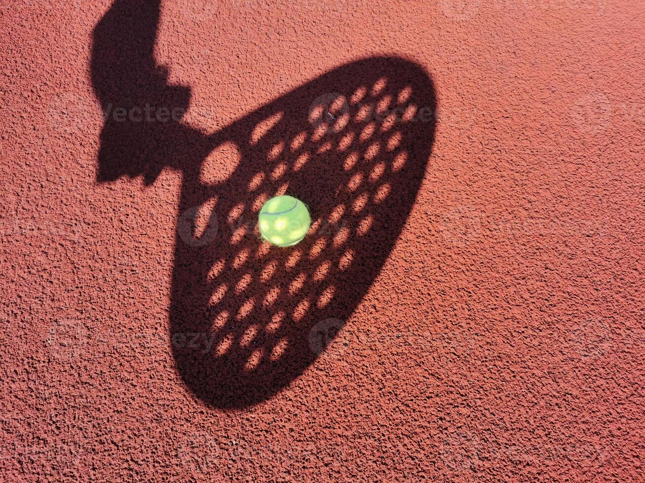 High angle of yellow ball placed on court and covered with shadow of crop person holding padel racket photo