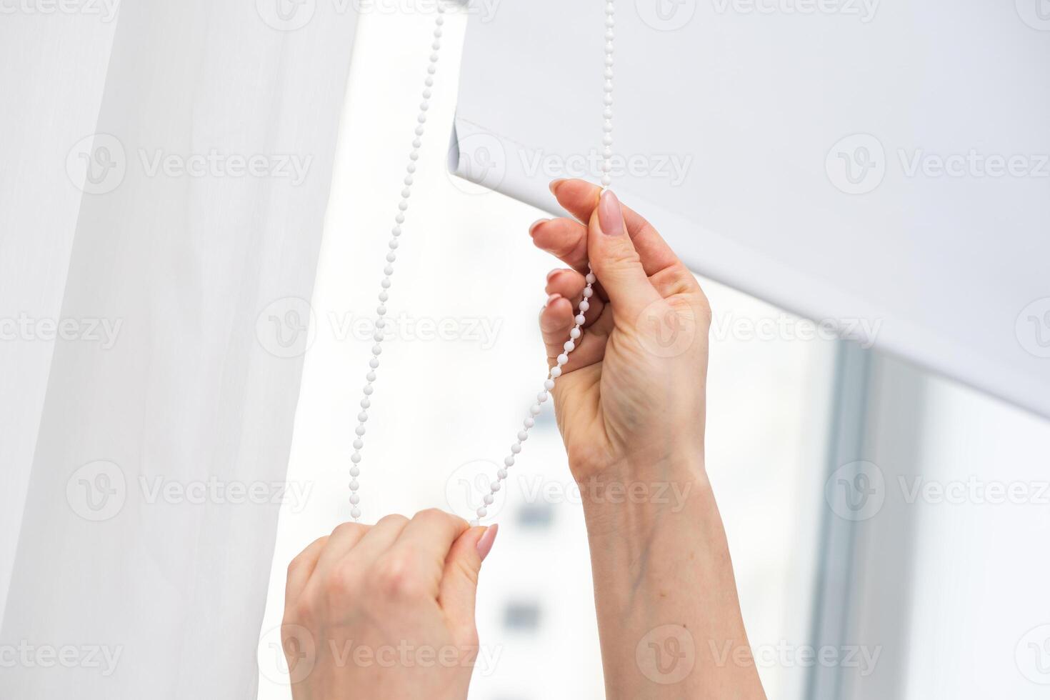 Woman opening white roller blind on window indoors, closeup photo