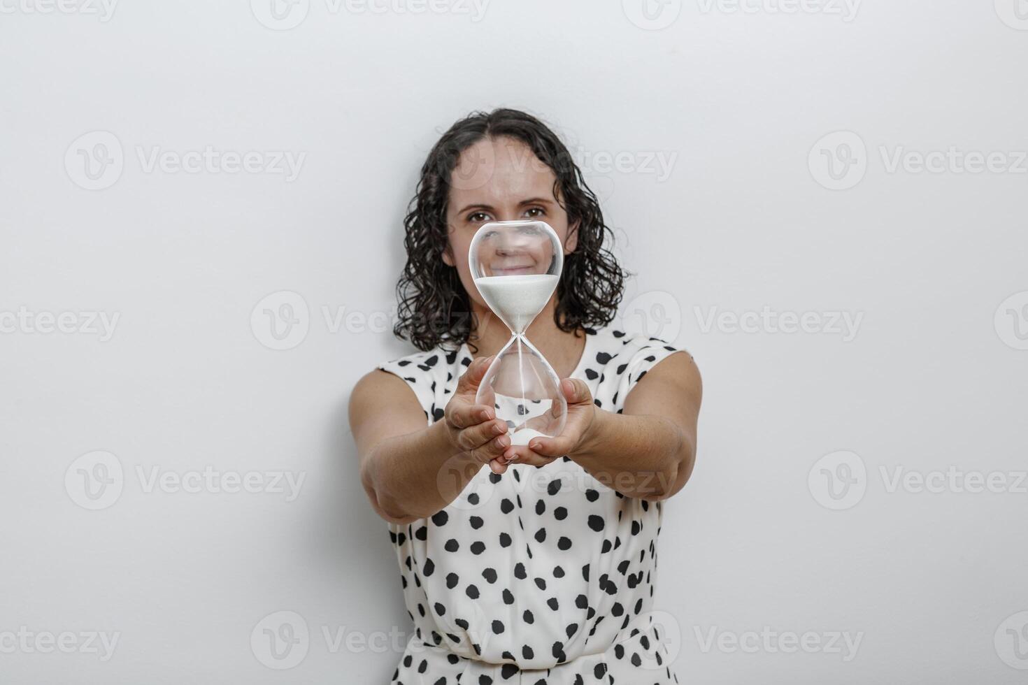 Woman in Polka Dot Dress Holding Transparent Hourglass With Sand Flowing at Studio photo