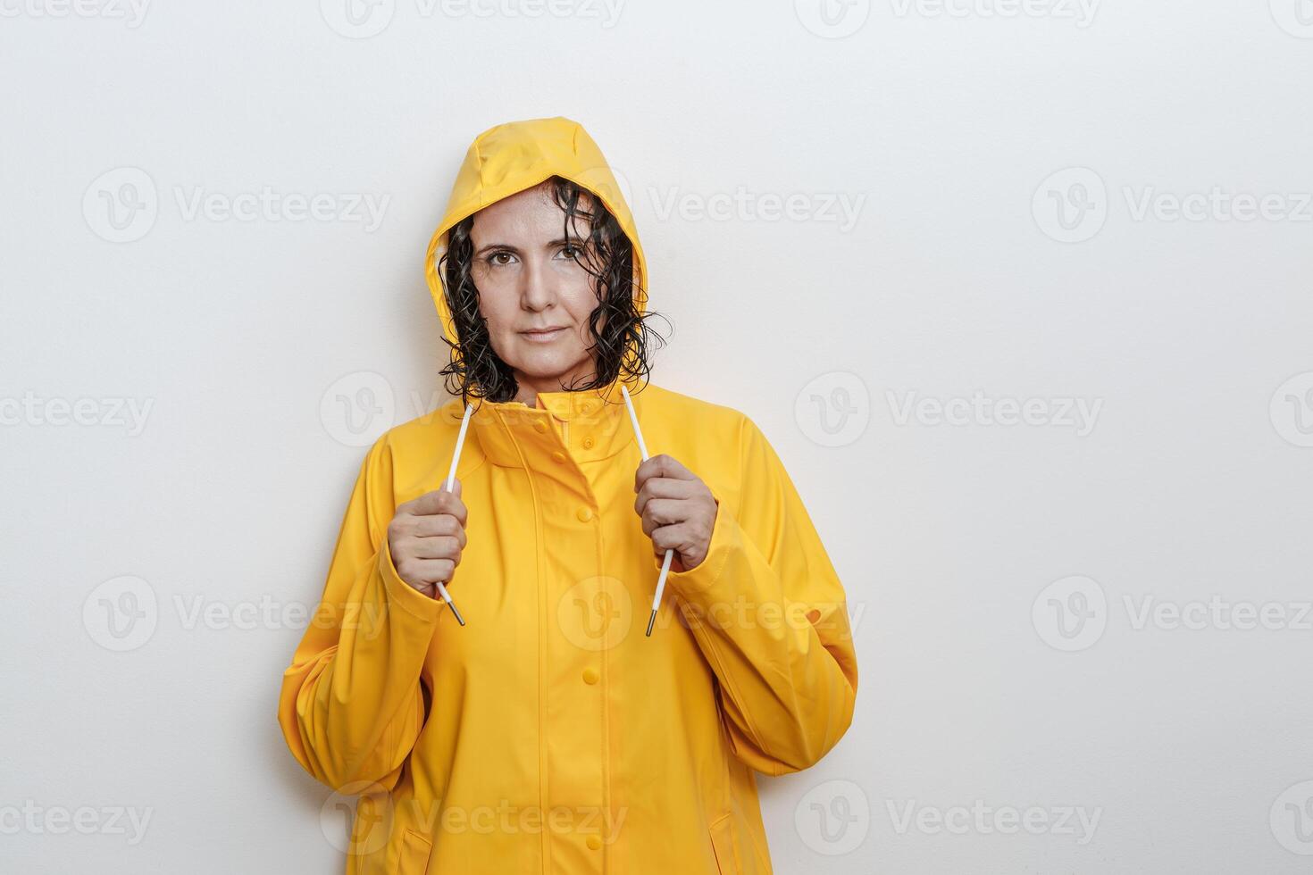 A woman with curly hair confidently stands against a white wall in a bright yellow rain jacket photo