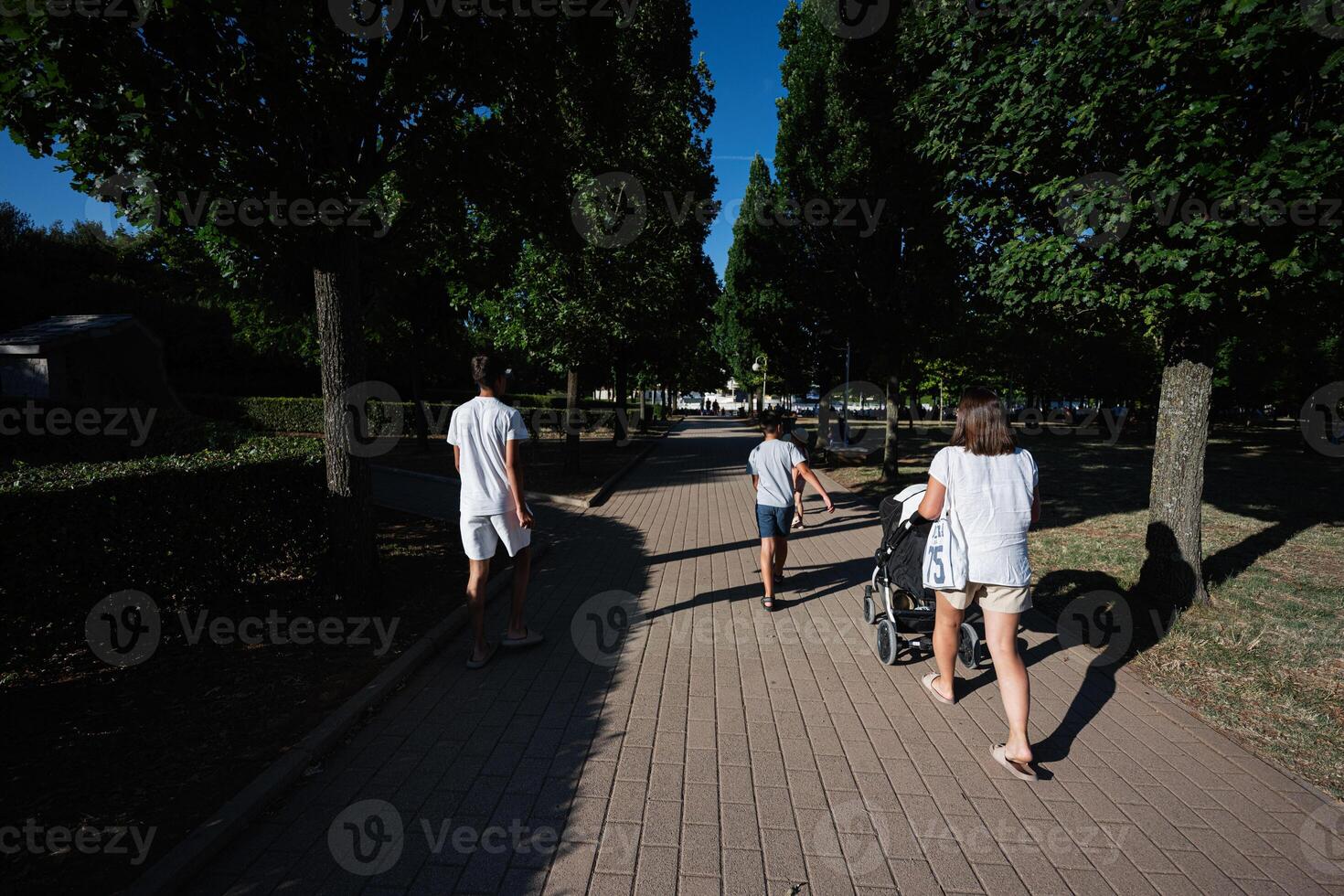 Family enjoying a walk in a park on a sunny day, with children and a stroller under the shade of tall green trees in Medjugorje. photo