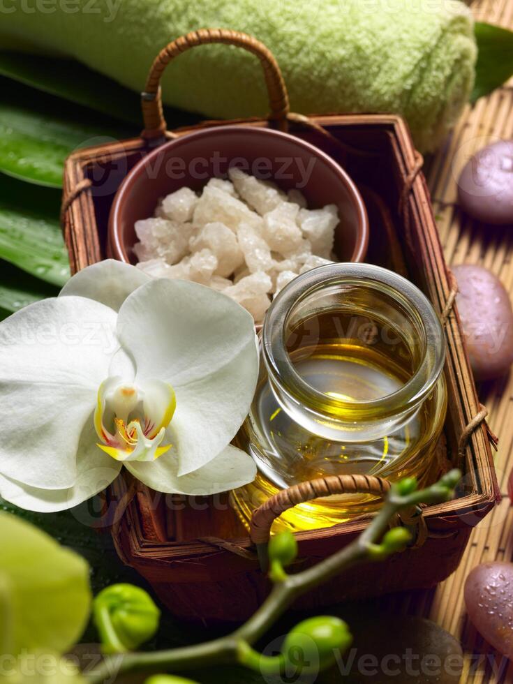 A tranquil spa setting featuring an orchid, essential oils, and rough sugar crystals, set against a backdrop of vibrant green leaves and a soft towel. photo