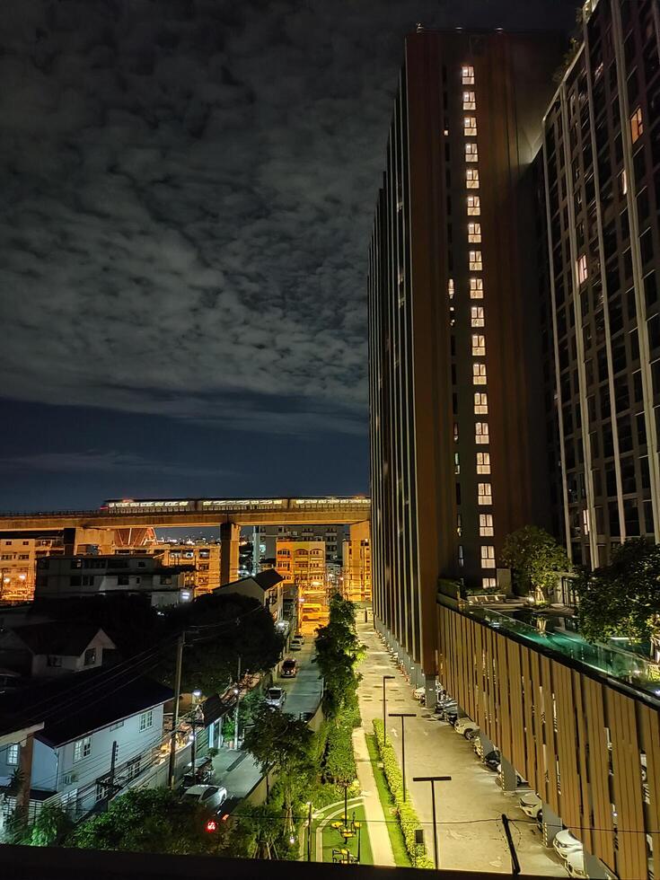 Image of a city with tall buildings on the side, a skytrain running and houses at night with neon lights. The sky is dark blue, small clouds fill the sky. photo