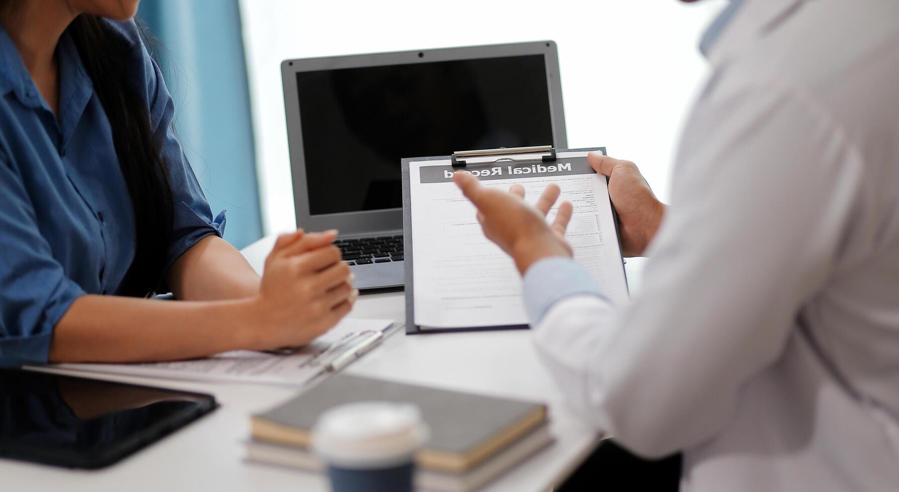Doctor and patient sitting near each other at the wooden desk in clinic. Female physician's pointing to a records form. Medicine concept photo