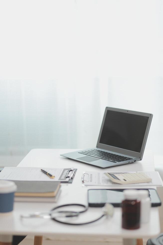 Doctor's working on laptop computer, writing prescription clipboard with record information paper folders on desk in hospital or clinic, Healthcare and medical concept. Focus on stethoscope photo