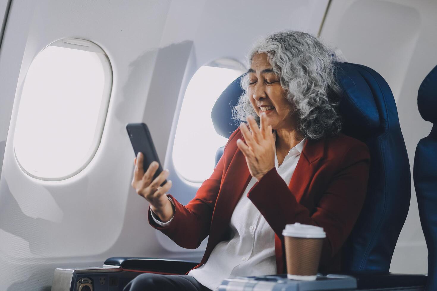 Portrait of a successful Asian businesswoman or entrepreneur in a formal suit on an airplane sitting in business class using a phone, computer laptop. Travel in style, work with grace. photo