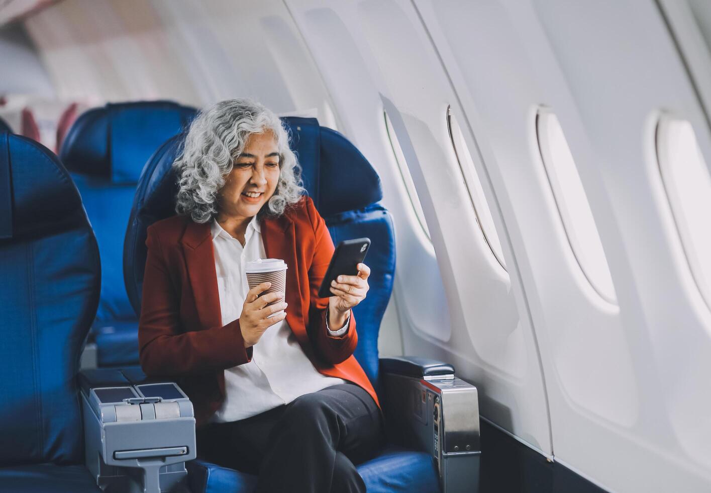 Portrait of a successful Asian businesswoman or entrepreneur in a formal suit on an airplane sitting in business class using a phone, computer laptop. Travel in style, work with grace. photo