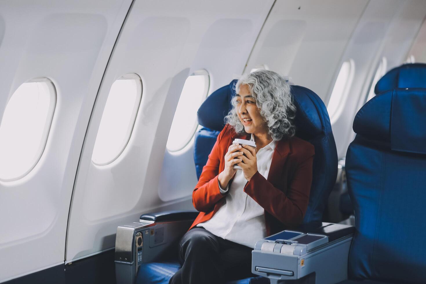 Portrait of a successful Asian businesswoman or entrepreneur in a formal suit on an airplane sitting in business class using a phone, computer laptop. Travel in style, work with grace. photo
