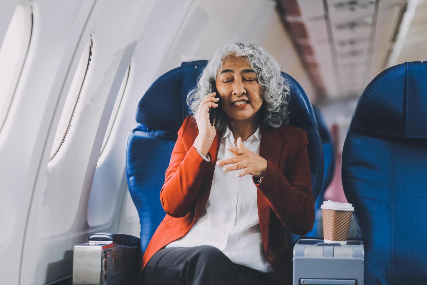 Portrait of a successful Asian businesswoman or entrepreneur in a formal suit on an airplane sitting in business class using a phone, computer laptop. Travel in style, work with grace. photo