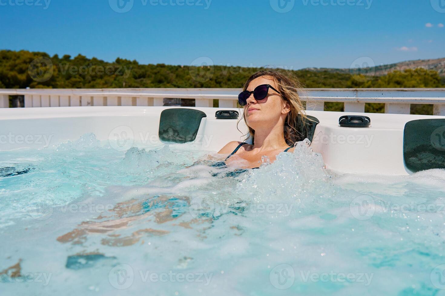 Woman wearing sunglasses and swimsuit relaxing in outdoor hot tub with bubbling water and enjoying sunny day. Female resting in outdoor jacuzzi with beautiful natural landscape photo