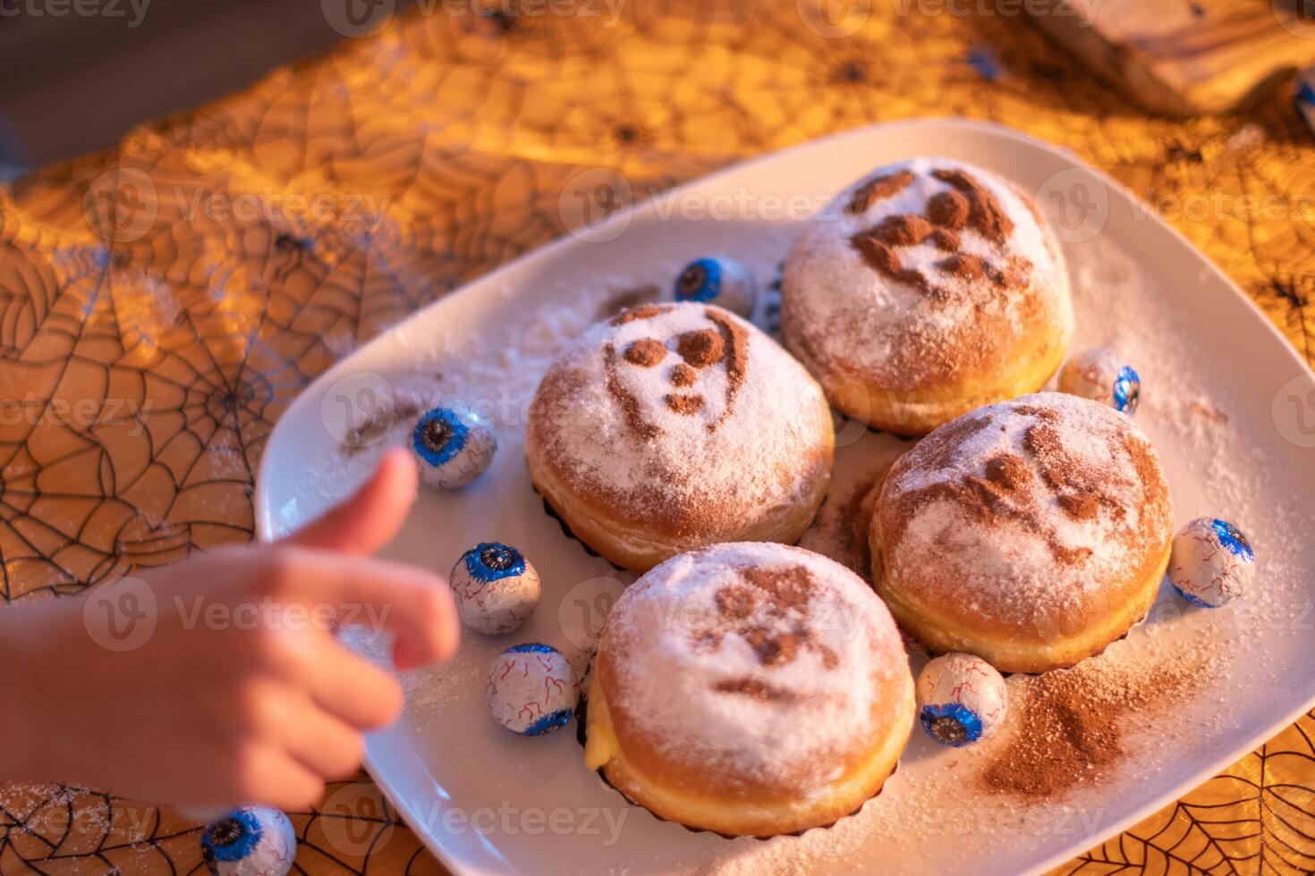 Child's hand reaching for bun, Four Halloween themed sweet buns with spooky faces sprinkled with powdered sugar on white plate, Spider web pattern background, Halloween dessert, photo