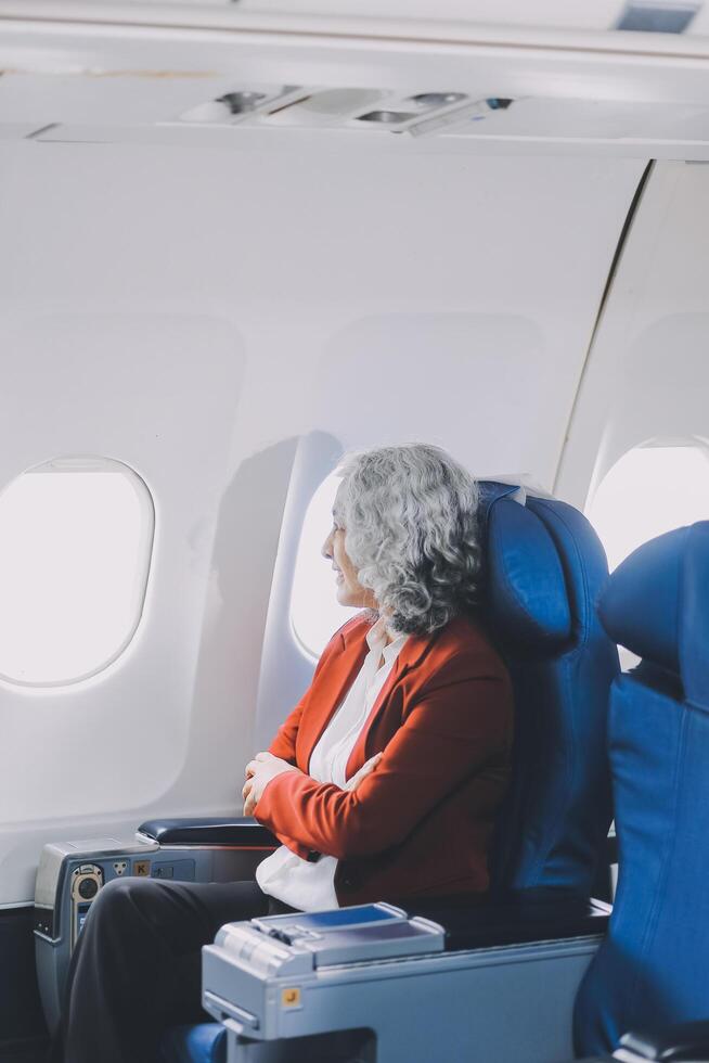 Portrait of a successful Asian businesswoman or entrepreneur in a formal suit on an airplane sitting in business class using a phone, computer laptop. Travel in style, work with grace. photo