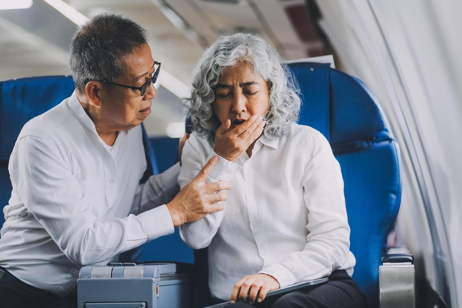 A mature woman sits by the window on a passenger plane, traveling abroad. She looks unwell, experiencing a headache, nausea, and vomiting, which dampens her travel excitement. photo