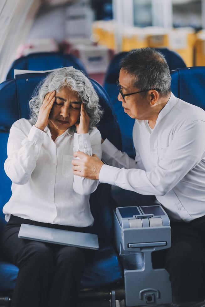 A mature woman sits by the window on a passenger plane, traveling abroad. She looks unwell, experiencing a headache, nausea, and vomiting, which dampens her travel excitement. photo