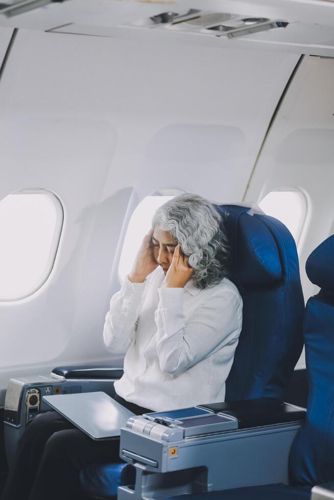 A mature woman sits by the window on a passenger plane, traveling abroad. She looks unwell, experiencing a headache, nausea, and vomiting, which dampens her travel excitement. photo