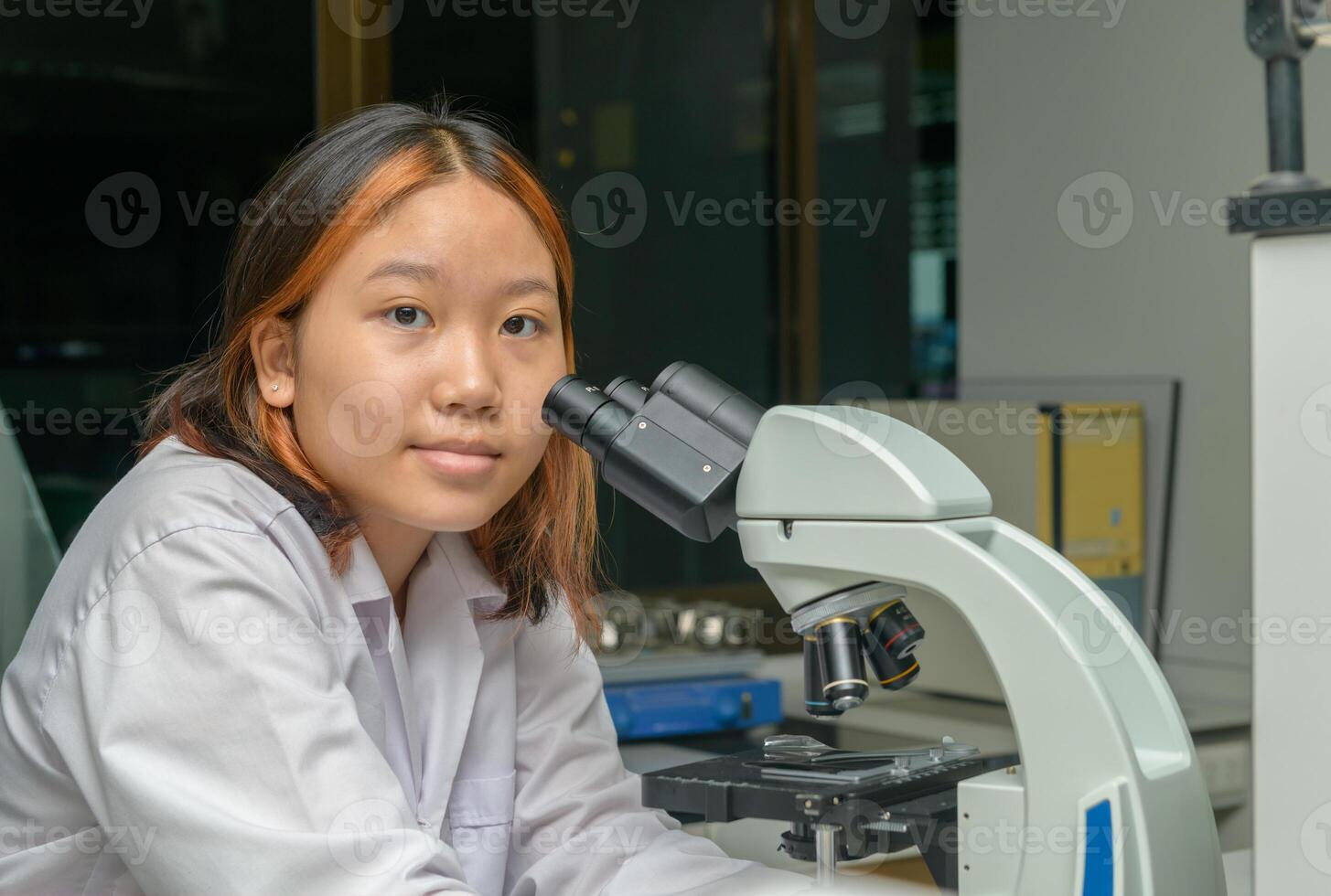 Cute scientist schoolgirl in lab coat looks through microscope for study microbiology in laboratory. photo