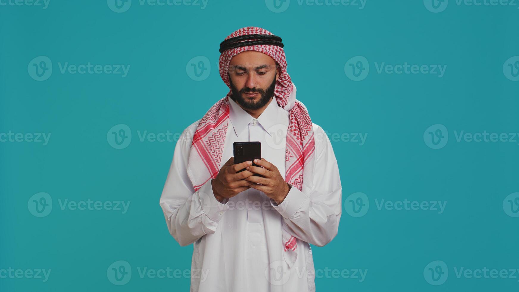 Islamic man works on his mobile gadget, surfing through social media platforms or online pages. Middle eastern guy in customary apparel uses smartphone to access global web, feeling excited. photo