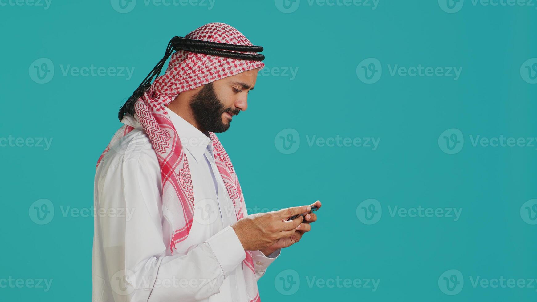 Islamic guy wearing traditional clothes playing online game on smartphone, standing against blue background. Arab gamer enjoying gaming tournament on mobile phone, entertainment. photo