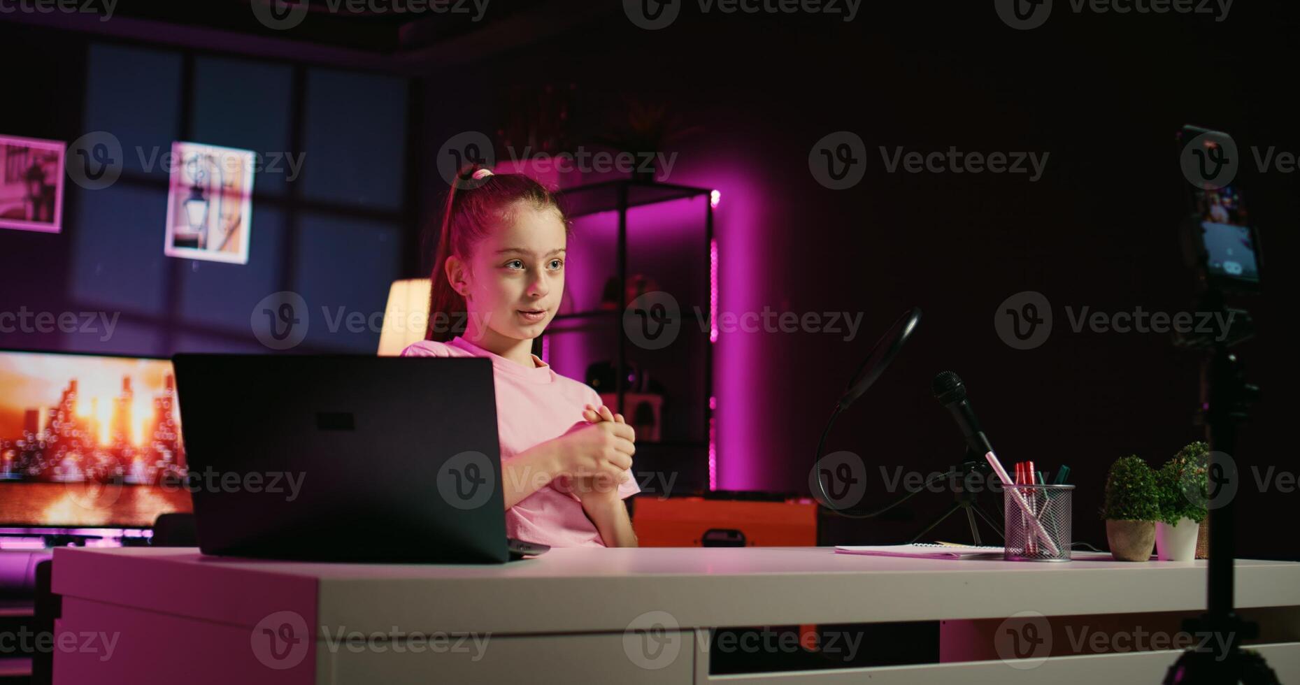 Dolly in shot of happy girl shooting for social media platform in living room, talking about her day. Joyous kid entertaining fans, doing content creation using phone on tripod photo