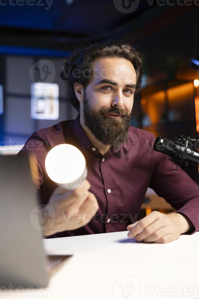 Smiling tech guru reviewing smart light bulb in studio, discussing with audience about home improvement. Cheerful man sharing latest news in WiFi home automation industry photo