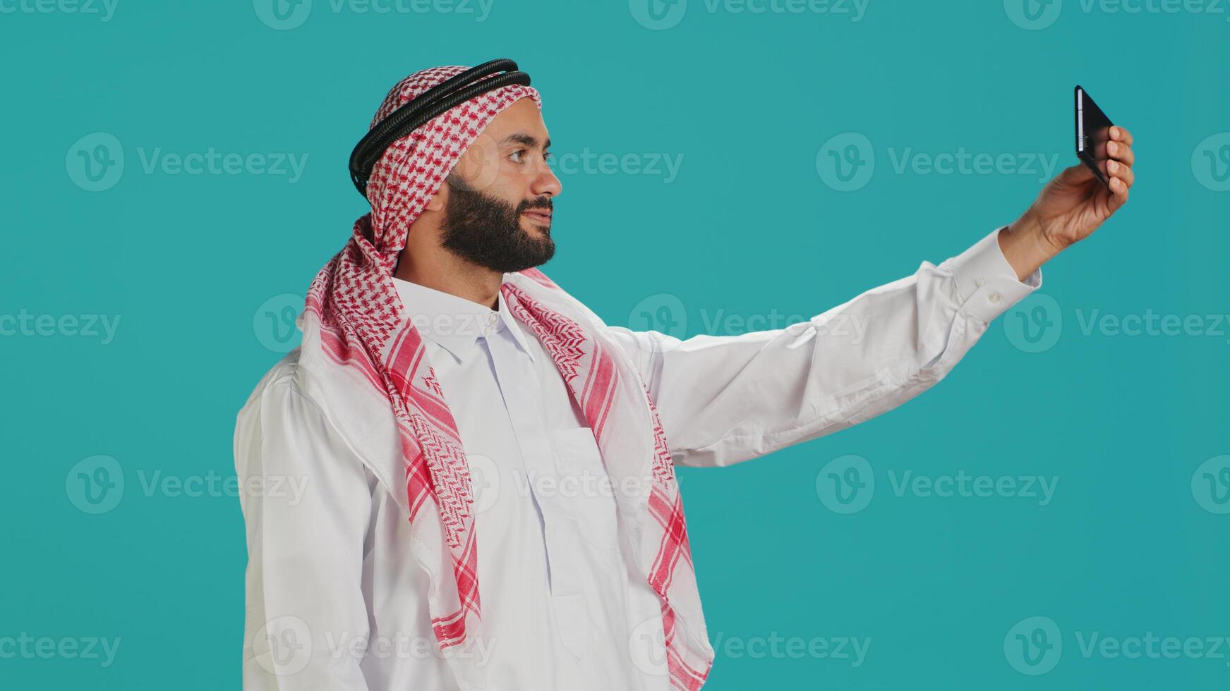 Young arab person takes photos with phone, capturing funny moments on smartphone camera in studio. Middle eastern guy with national headscarf taking pictures in traditional attire.