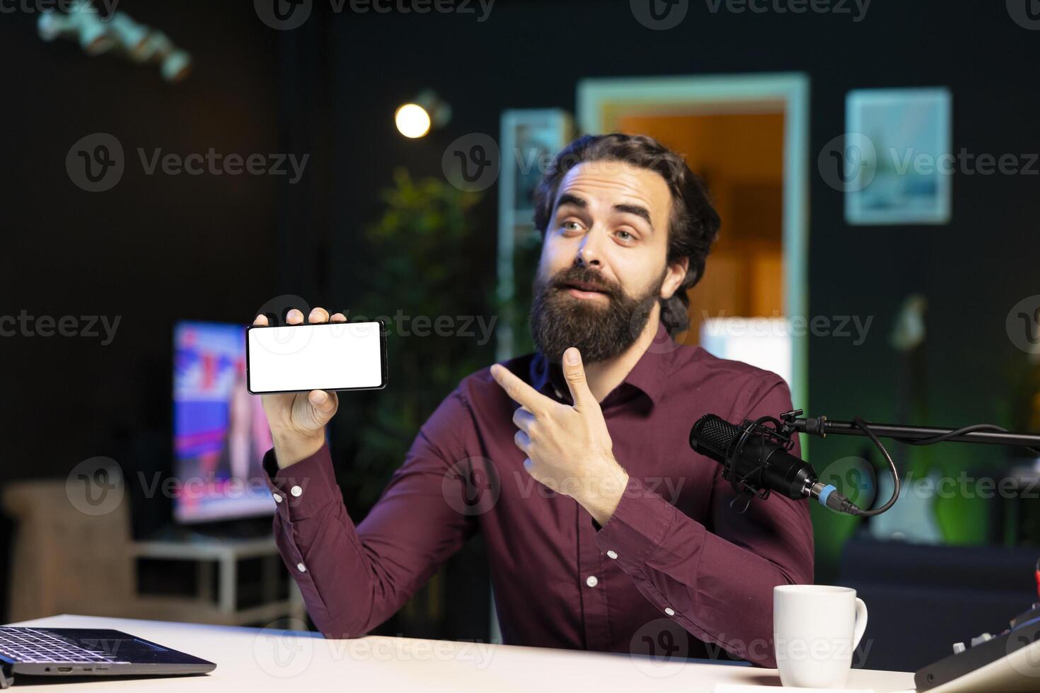 Man pointing at isolated screen cellphone in hand, using it as empty copy space to showcase his online streaming channel logo. Joyful media star does introductory using mockup phone photo