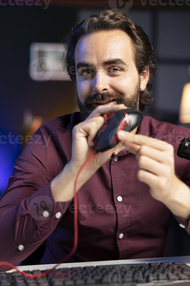 Portrait of cheerful influencer filming for online channel in apartment studio, presenting gaming mouse to audience. Joyful man showing viewers computer peripheral device, recommending it photo