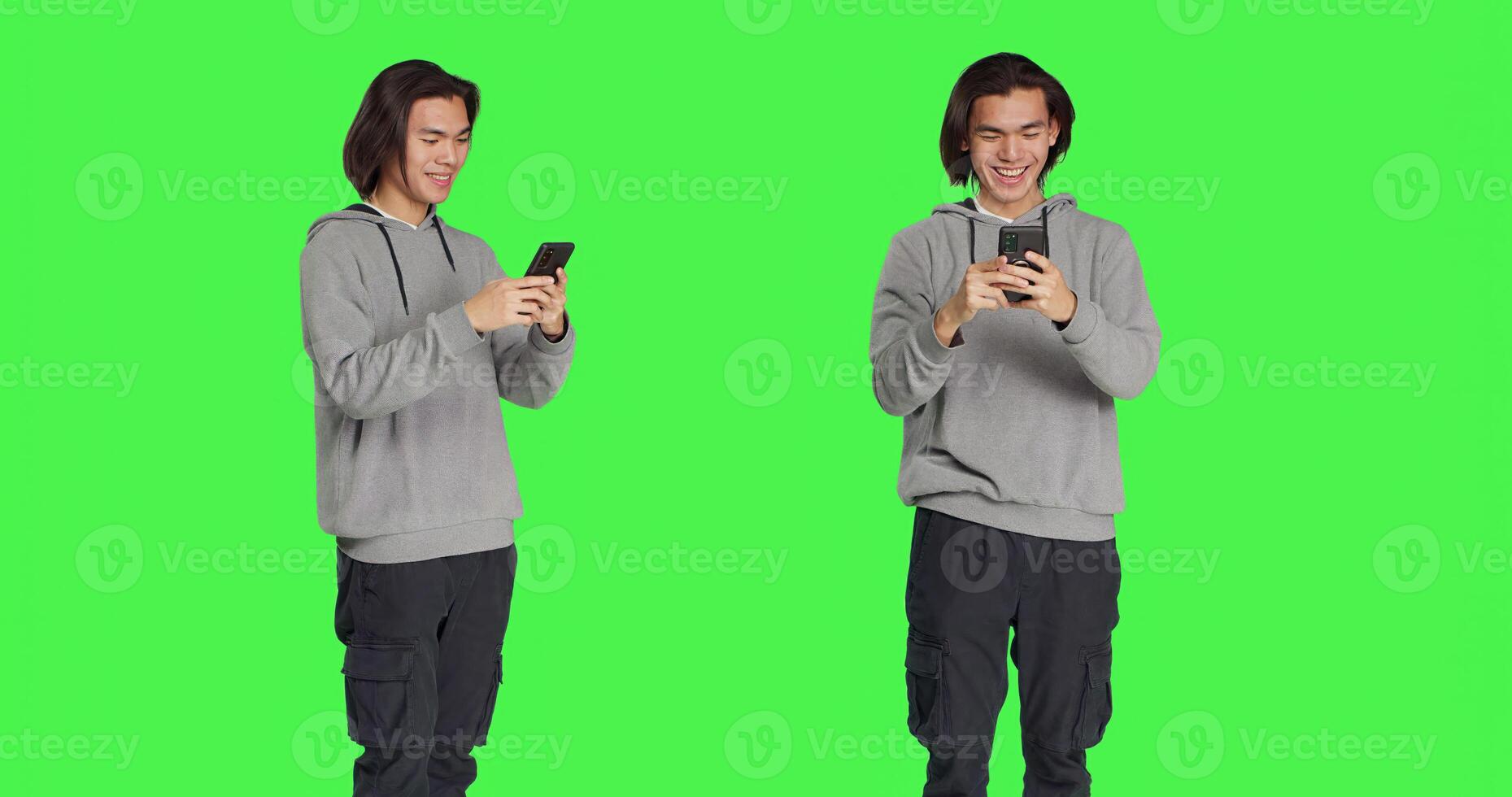 Guy texting messages on mobile phone standing against greenscreen backdrop, using social media app to chat remotely with friends. Asian joyful adult messaging on smartphone. photo
