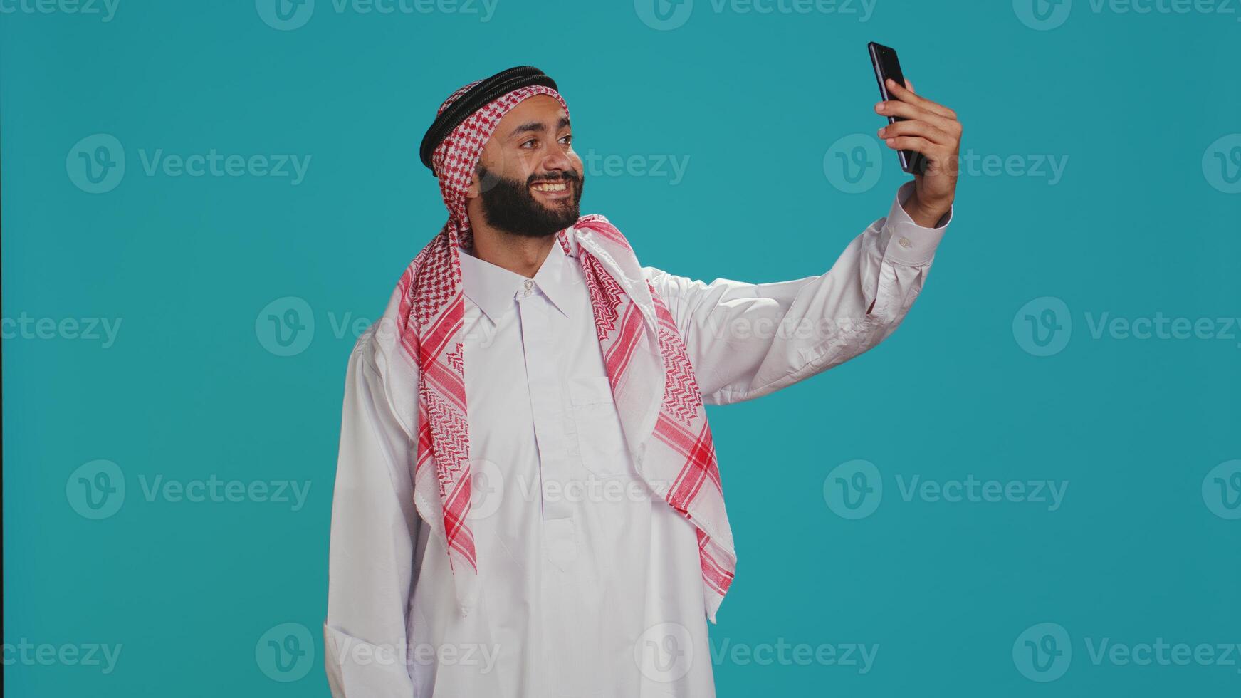 Young adult taking photos with phone, capturing memories and funny faces on smartphone camera over blue background. Middle eastern guy with checkered headscarf takes pictures in traditional attire.