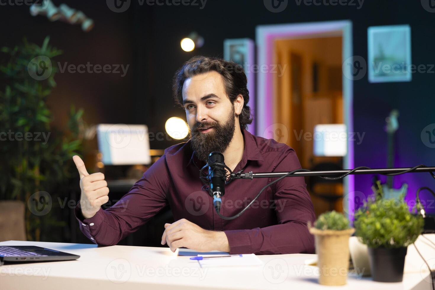 Content creator hosting online show in rgb lights studio, shooting vlog using professional microphone, showing thumbs up sign. Vlogger doing hand gesturing during streaming service broadcast photo