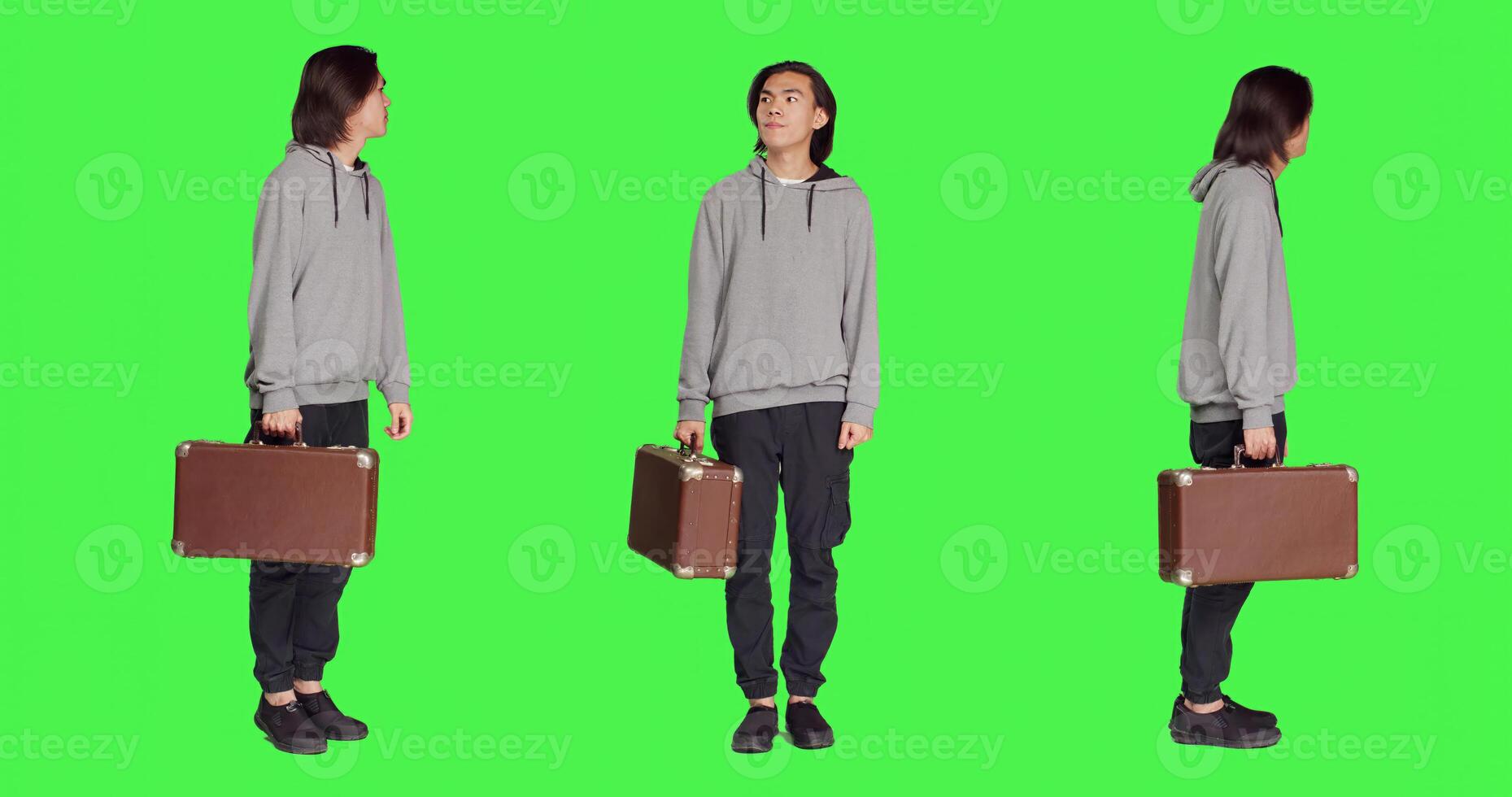 Young adult carries retro suitcase over full body greenscreen backdrop on camera, waiting impatiently for a thing. Asian model holding big suitcase while he checks time on wrist watch. photo