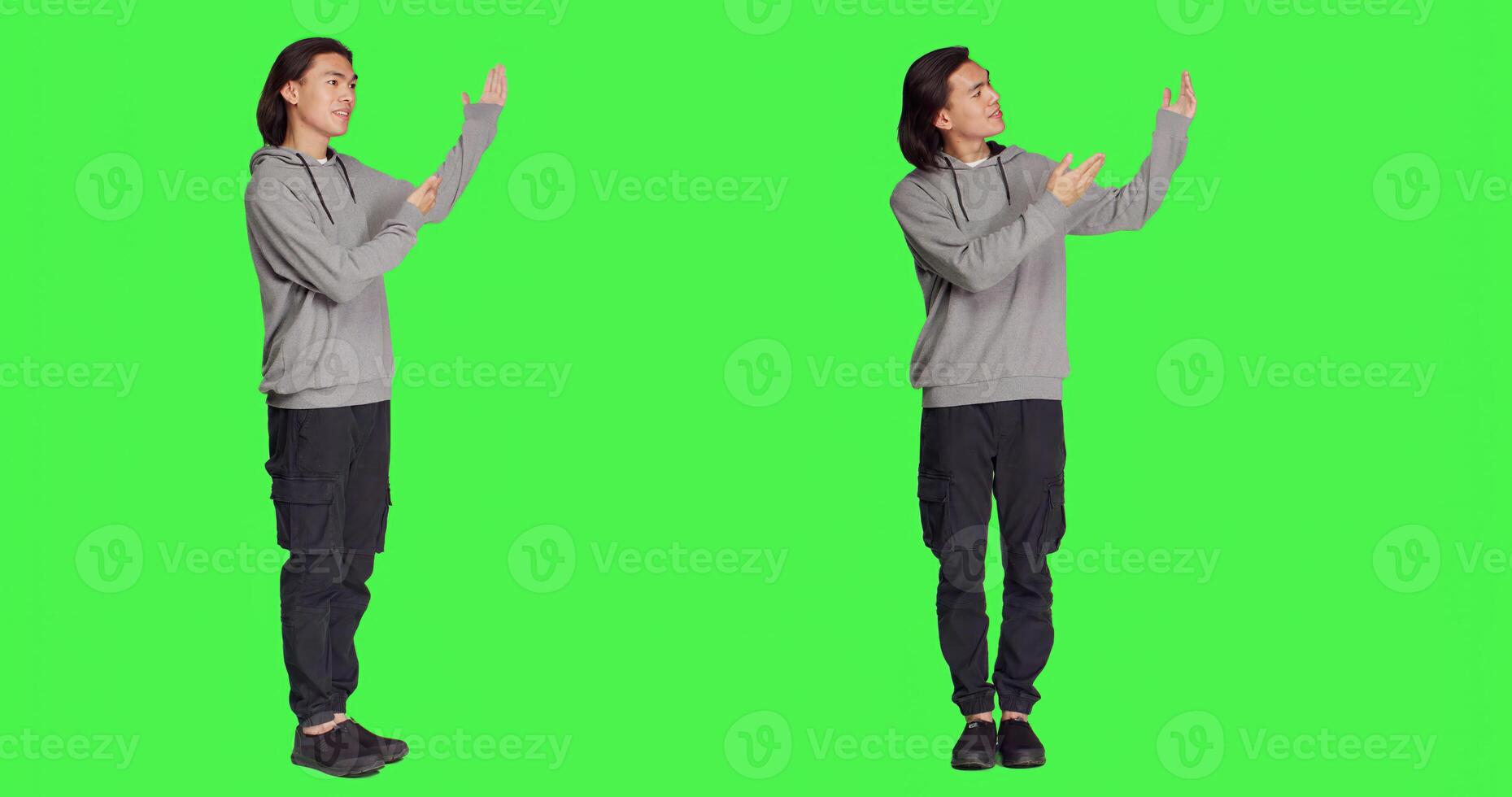 Asian man doing advertisement in studio, pointing at something next to him for new ad slogan. Young person smiling and working on promotional marketing, standing over full body greenscreen backdrop. photo