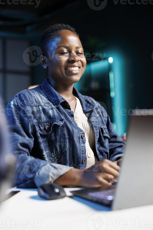 Close-up of African American lady smiling enthusiastically while typing on a digital laptop. Joyful black woman sitting at desk and using personal computer, researching and reviewing emails. photo