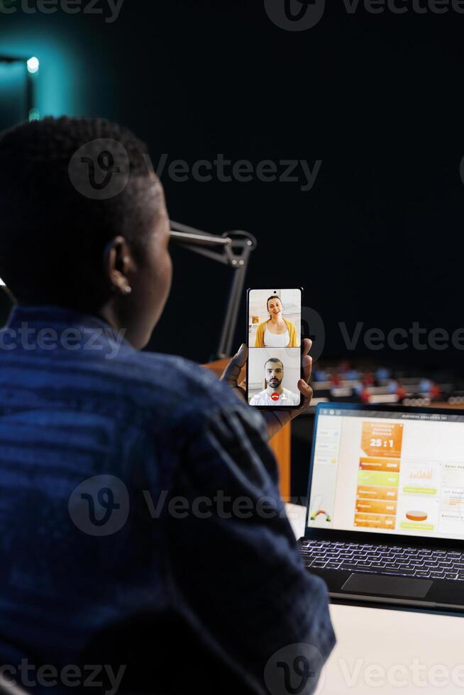 Over the shoulder shot of freelancer using mobile device to communicate with others. African american woman using her cellphone to make a conference call while sitting at a table with laptop. photo