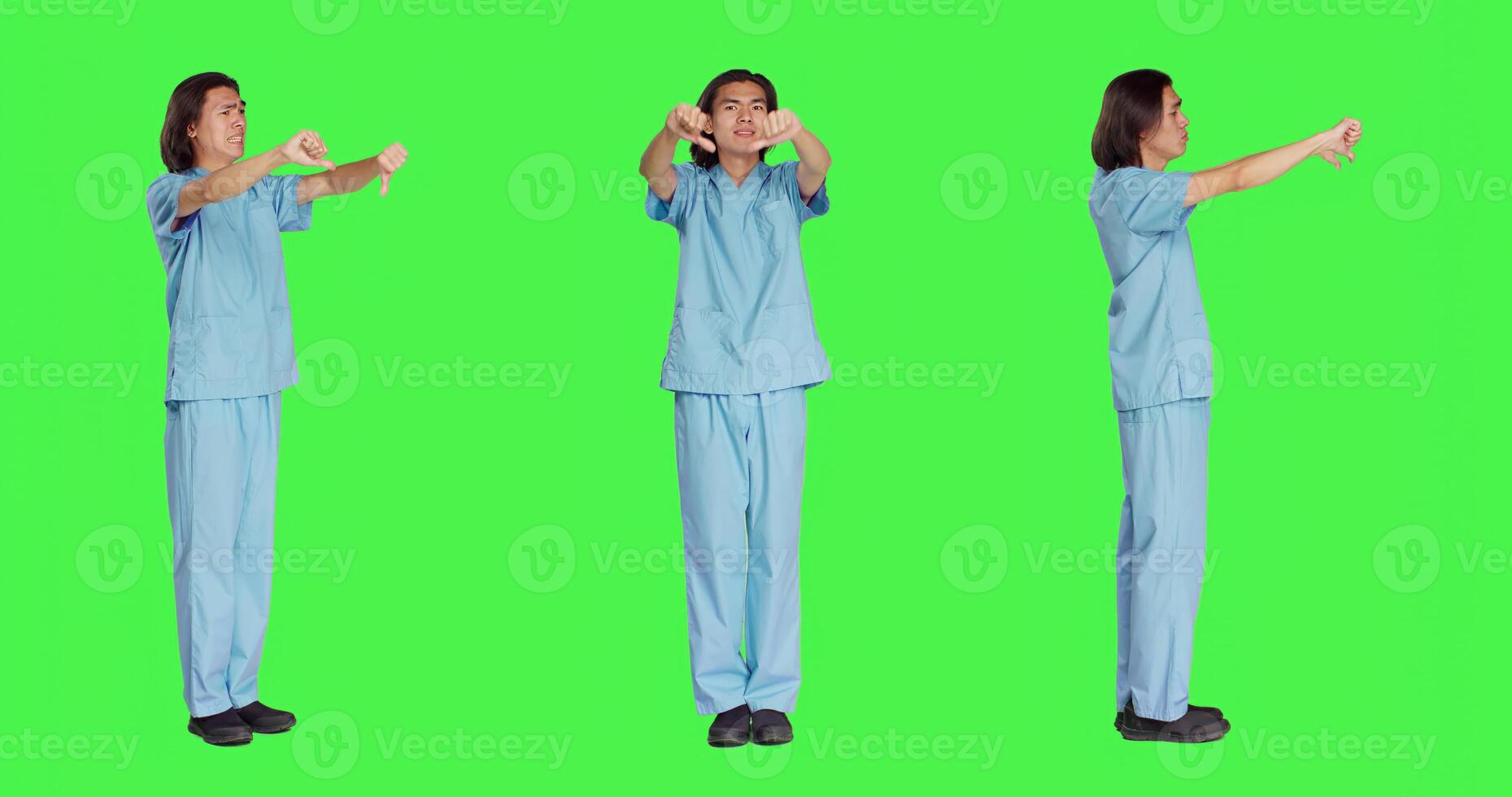 Asian nurse giving thumbs down in studio, showing dislike and negativity symbol over full body greenscreen backdrop. Displeased medical assistant expressing disagreement and disapproval. photo