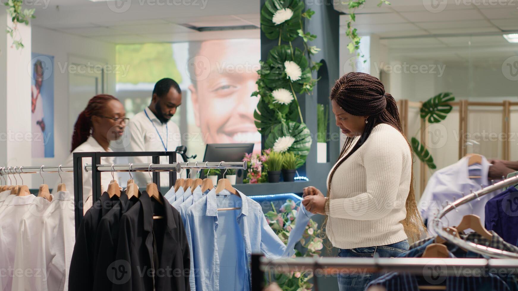 Woman doing shopping in elegant fashion boutique, looking at clothes on hangers. BIPOC client checking shirt materials quality from new collection stock of trendy garments on racks photo