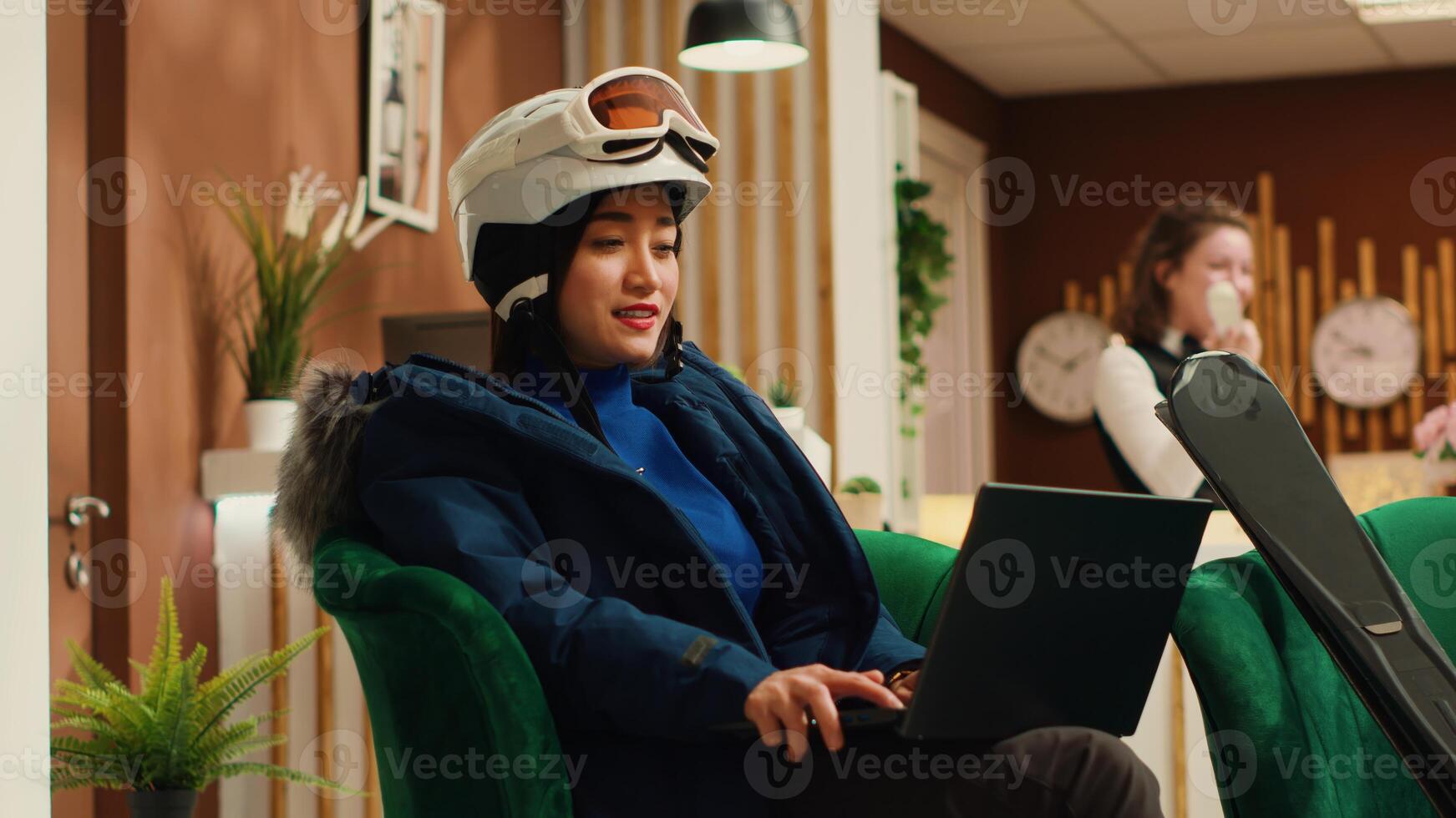 Asian woman working on laptop while on wintertime vacation at mountain resort, sending emails before going down slopes with skis gear. Hotel guest wearing snow suit and helmet. Handheld shot. photo