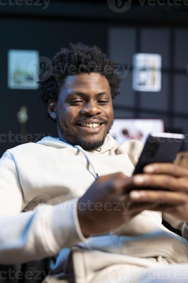 Cheerful man impatiently texting friend, discussing meeting hour. Person sitting on cozy sofa in modern stylish apartment typing on mobile phone, excited to meet up with mate photo