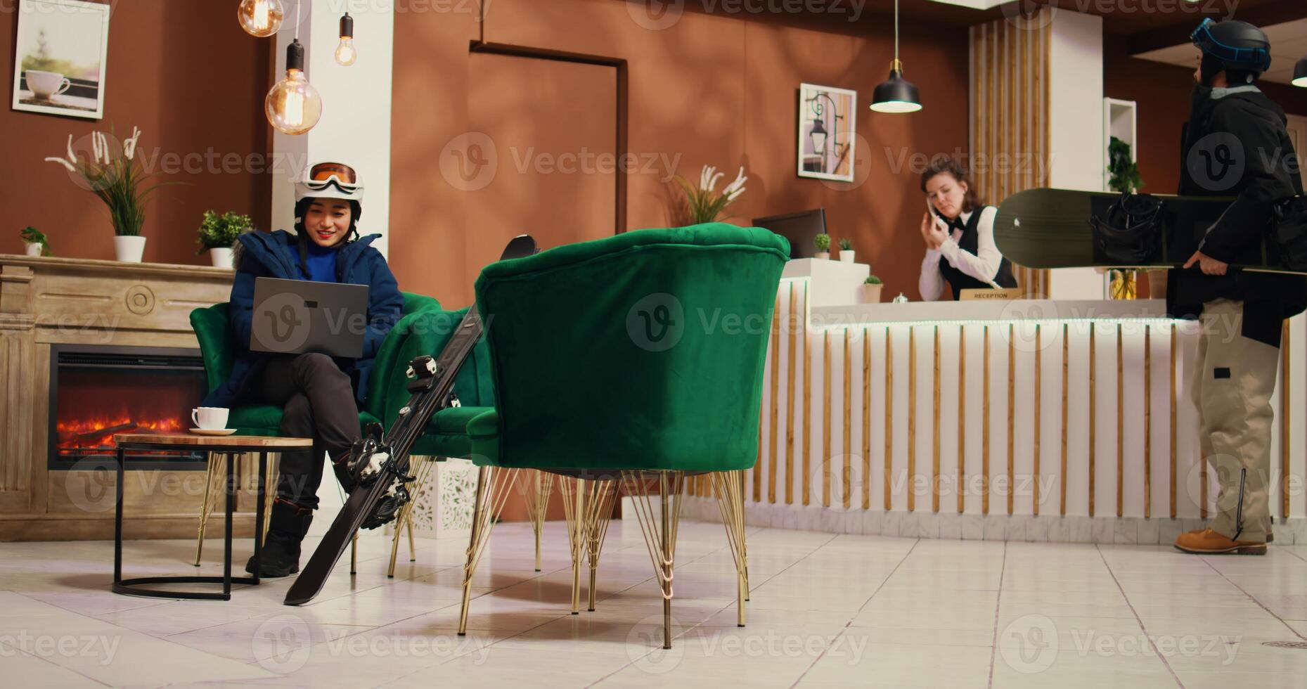 Asian person working on laptop in lobby while on wintertime holiday at mountain alpine resort, sending emails before going down slopes with skis. Guest wearing snow attire and helmet. photo
