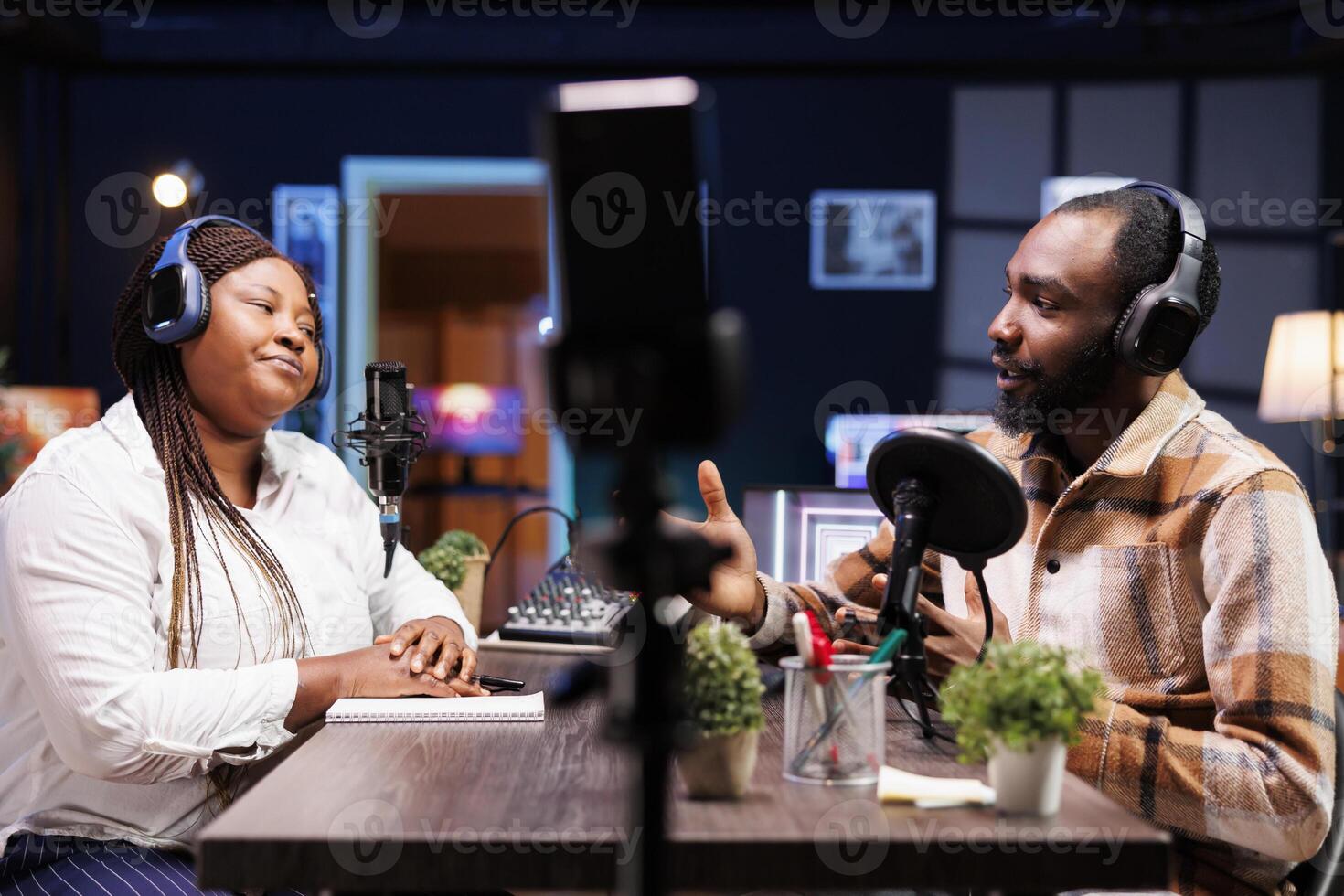 Cohosts filming podcast episode using smartphone on tripod, broadcasting on vlog channel. African american man and woman meeting in studio to record show with mobile phone and talk politics. photo